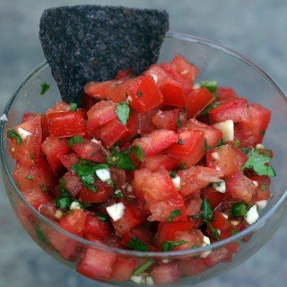Fresh pico de gallo in a glass bowl with blue corn tortilla being dipped into the salsa