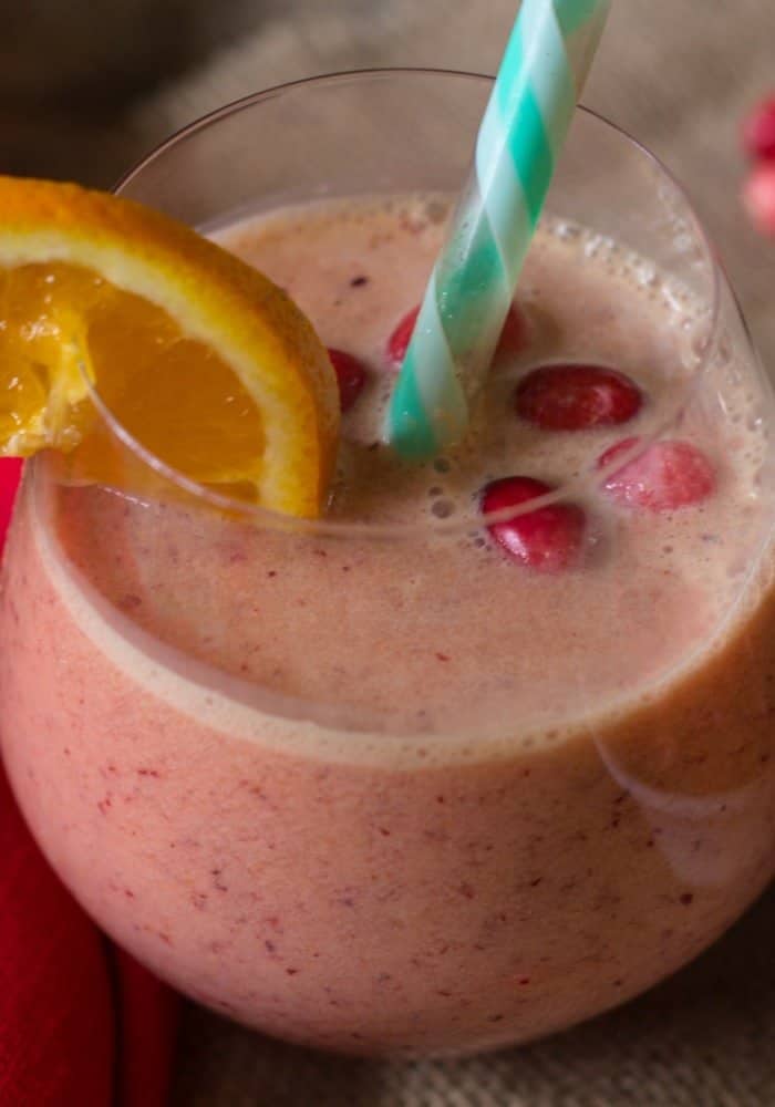 cranberry smoothie in a clear glass with a green and white straw and garnished with an orange and fresh cranberries.