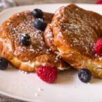 Berries and cream cheese french toast on a white plate topped with fresh berries and warm maple syrup. Sprinkled with confectioners sugar