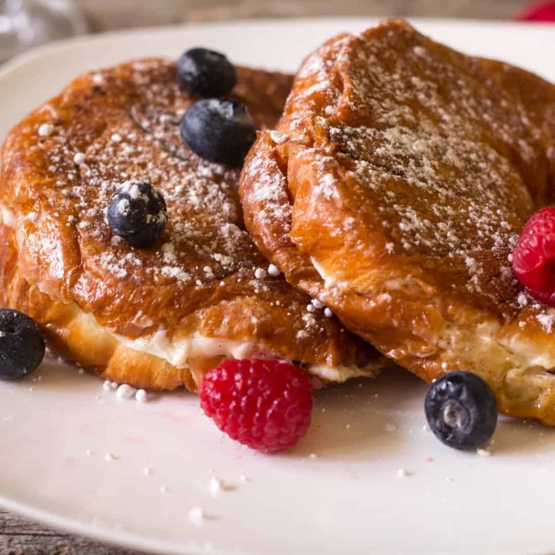 Berries and cream cheese french toast on a white plate topped with fresh berries and warm maple syrup. Sprinkled with confectioners sugar