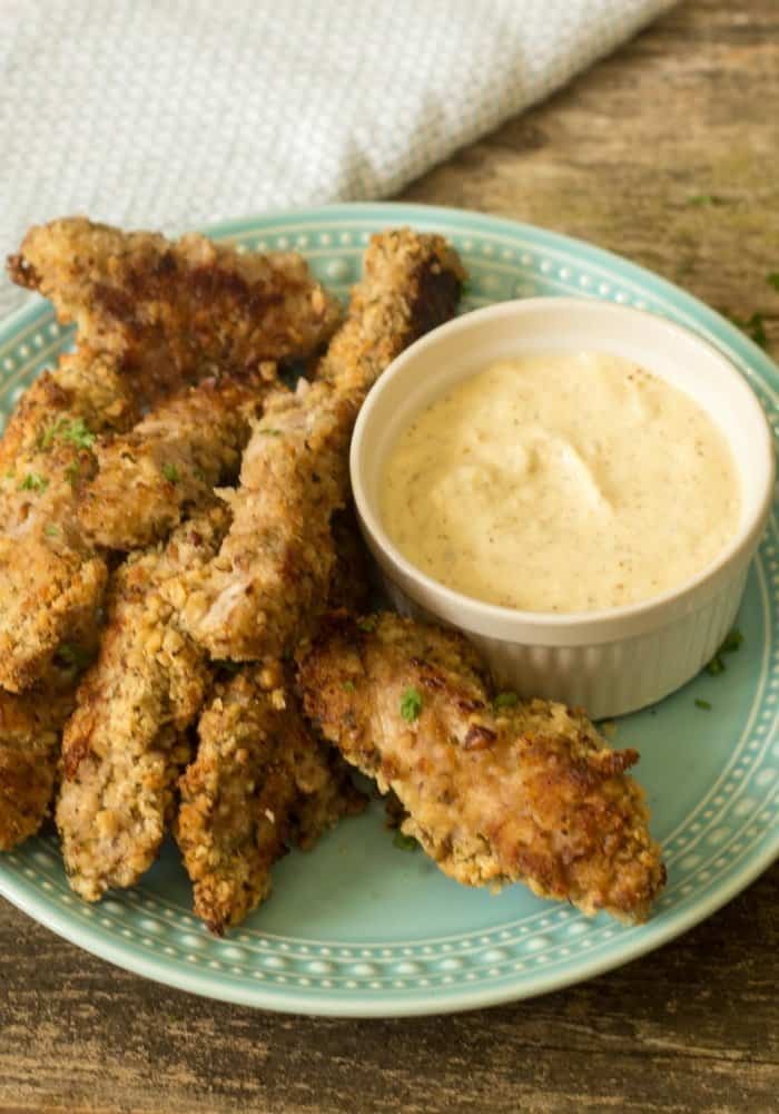 Side view of pecan crusted chicken tenders on a sky blue antique plate with a floral edge and a side sauce of creole dipping sauce in a white dipping bowl 