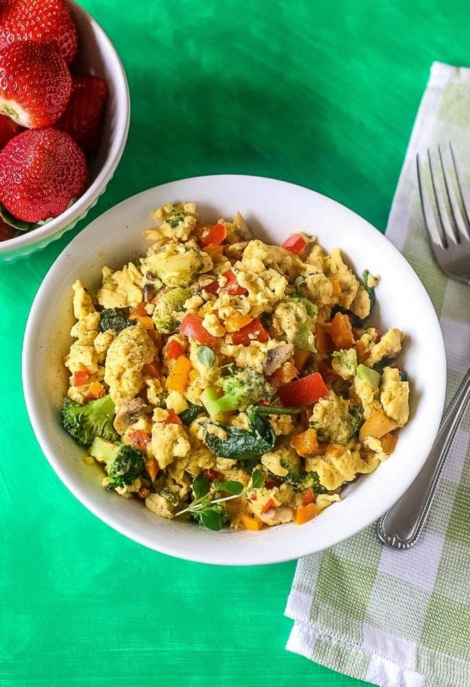 An aerial view of eggs scrambled with healthy veggies like broccoli, spinach and tomatoes in a white bowl with fresh strawberries in a side bowl