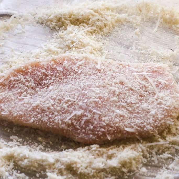 chicken breasts coated with almond flour and Parmesean cheese which on a white cutting board. The cutlets will be sauteed in light butter for chicken piccata