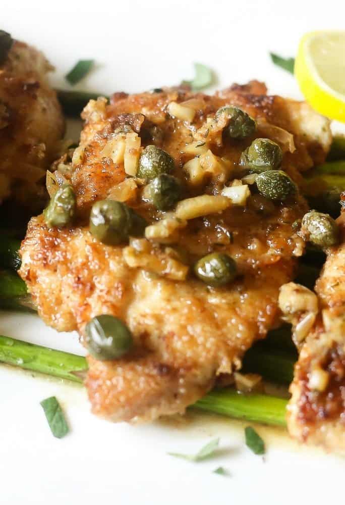 A side view of low carb lemon chicken piccata on roasted asparagus,The lemon chicken is topped with white wince sauce,capers,shallots on a white platter