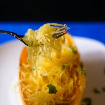 A side view of baked healthy spaghetti squash on a fork and in the background the squash is on a white platter with a bright blue background.