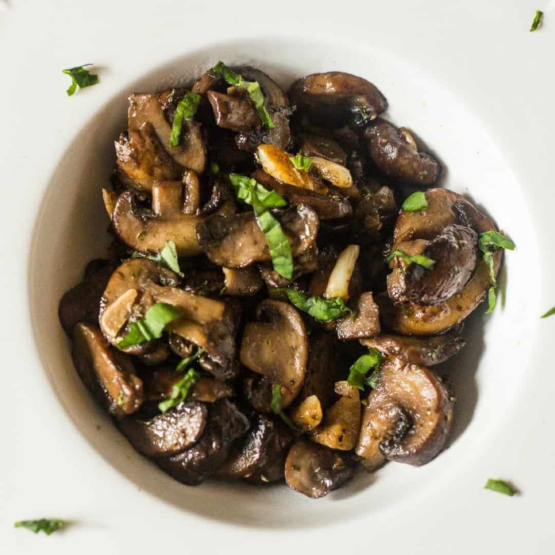 An aerial view of sliced sauteed mushrooms with garlic and wine with chopped parsley in a small white bowl