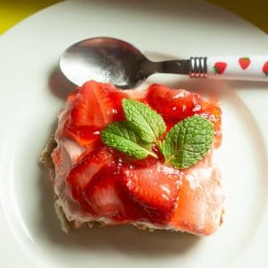 overhead view of strawberry pretzel jello recipe with mint leaves on top on a white dessert plate with a spoon on top of plate with strawberries on the handle