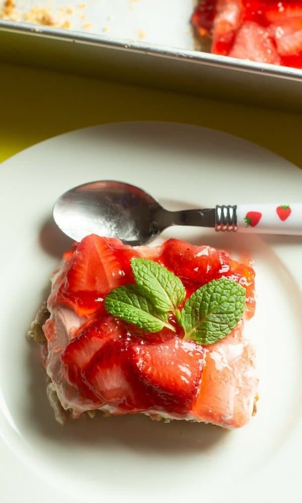 An overhead view of a slice of strawberry pretzel jello dessert on a white dessert plate and a spoon sitting above the slive with a strawberry decorated handle.