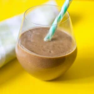 A side view of a chocolate protein smoothie with a green and white straw with a yellow backdrop.