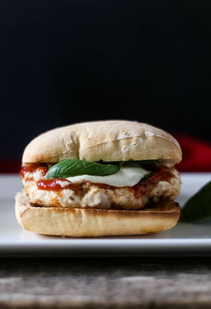 Sideview of a Parmesan chicken burger on a ciabatta roll, topped with homemade marinara, fresh basil leaves and melted fresh mozzarella.