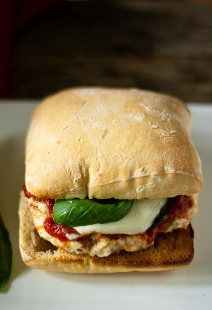 A side view of a grilled chicken parm burger with melted fresh mozzarella cheese and fresh basil on a toasted ciabatta roll. Served on a white rectangular plate.