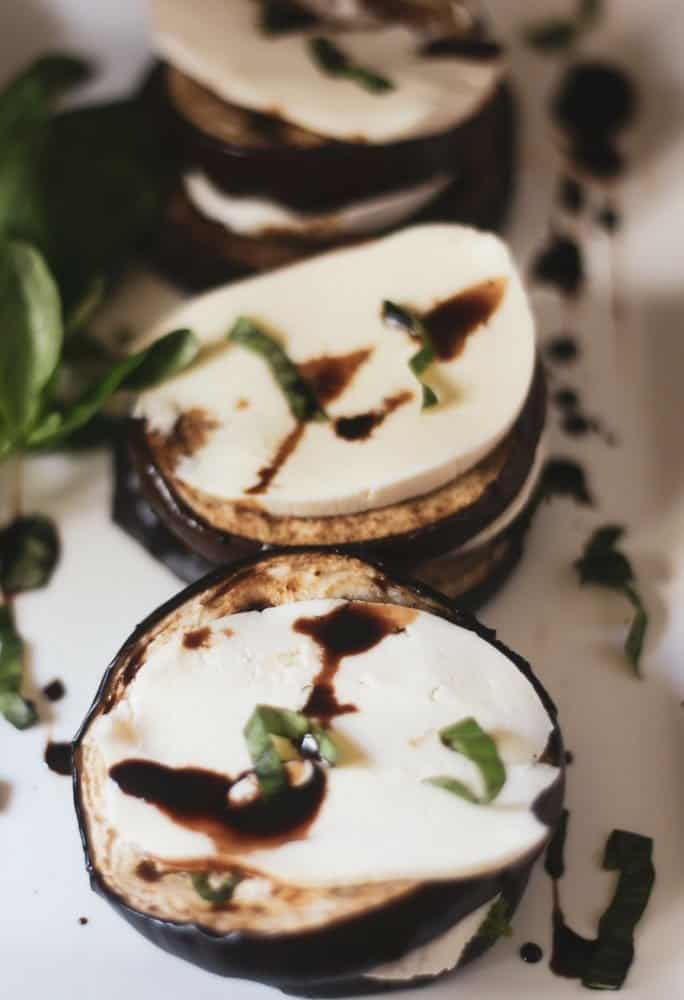An eggplant, tomato, mozzarella stack with basil and a balsamic glaze on a white platter.