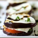A side view of a tasty and juicy eggplant Caprese salad layered with grilled eggplant, juicy vine ripe tomatoes, basil, fresh mozzarella and a balsamic glaze topping it and dripping down the side of the vegetable stack