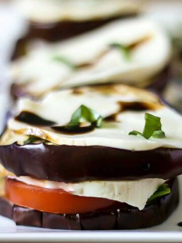 A side view of a tasty and juicy eggplant Caprese salad layered with grilled eggplant, juicy vine ripe tomatoes, basil, fresh mozzarella and a balsamic glaze topping it and dripping down the side of the vegetable stack