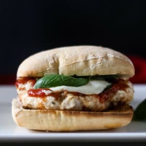 A side view of grilled chicken parm burger on a toasted ciabatta roll with homemade marinara sauce , fresh basil leaves and melted mozzarella.