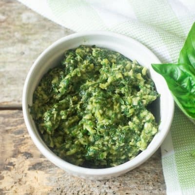 Skinny basil pesto in a white serving bowl loaded with garlic and Parmesan and fresh basil leaves to the right of the bowl on a checkerboard green and white linen.
