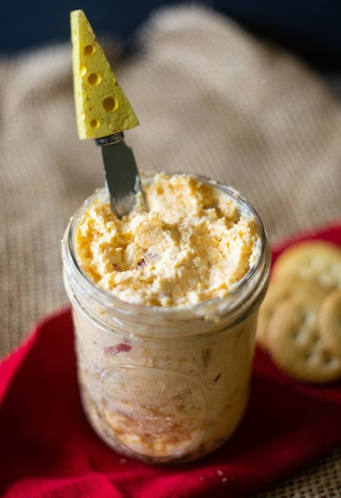Pimento Cheese dip in a mason jar along with a cheese spreader in the dip with a Swiss cheese head ready to be spread on crackers.