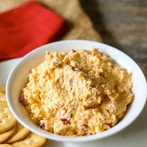 Side view of super creamy homemade pimento cheese dip in a white bowl with Ritz crackers and a red linen napkin in the background