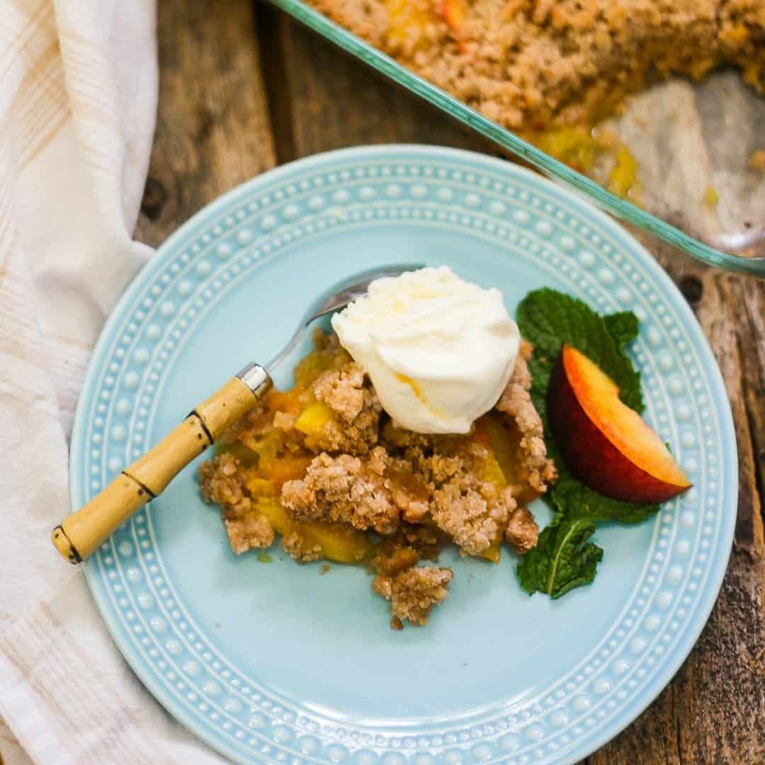 An overhead view of the most delicious peach crumble on a blue dessert plate topped with melting vanilla bean ice cream.