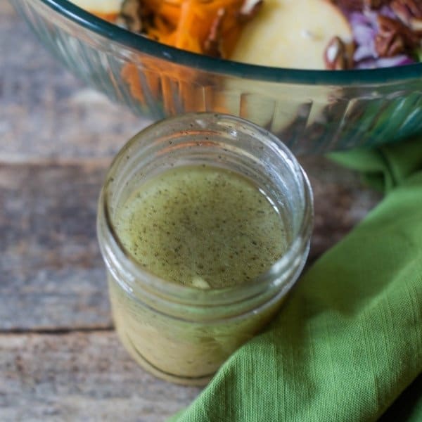 Maple Cider Vinaigrette in a mason jar with a green linen to the right of the jar and an apple pecan salad in a glass bowl behind the mason jar