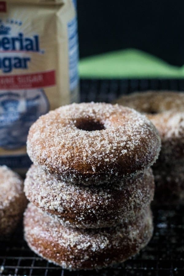 Three Baked Apple Cider Donuts with cinnamon sugar stacked on top of each other on a wire cooling rack with a bag of Imperial Sugar behind the donuts.