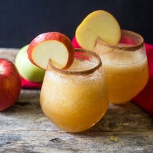 Two glasses of caramel apple vodka slush with the glasses rimmed with cinnamon sugar for garnish on a wooden table with apples a a red linen on the table.
