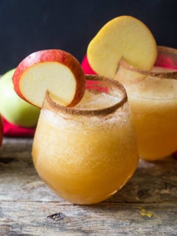 Two glasses of caramel apple vodka slush with the glasses rimmed with cinnamon sugar for garnish on a wooden table with apples a a red linen on the table.