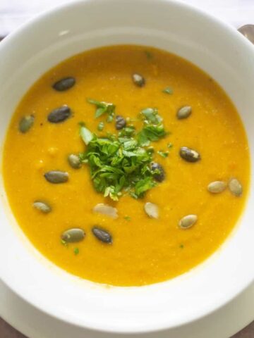 Hearty carrot coconut soup in a white soup bowl topped with pepitas and chopped herbs