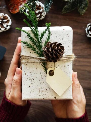 A gift wrapped with pretty white paper,a ribbon, green pine,and a pinecone.