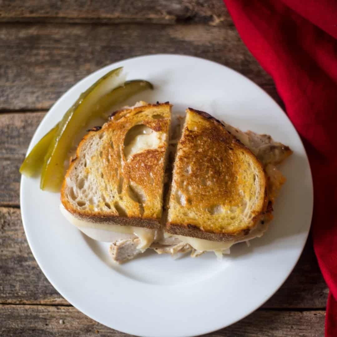 A delicious grilled golden brown crispy reuben sandwich with melted swiss cheese, Russian dressing, sauerkraut on a white plate with pickles.