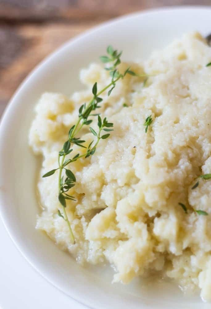 Mashed cauliflower in a white serving bowl with a sprig of thyme on top!