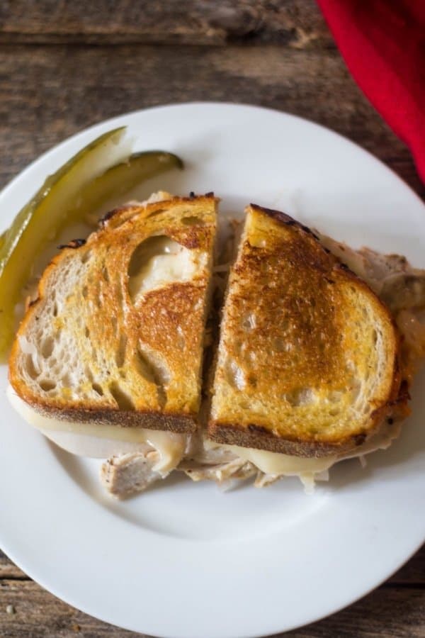 Turkey reuben sandwich which crispy golden rye bread, melted swiss cheese, and leftover roasted turkey on a white plate with pickles.