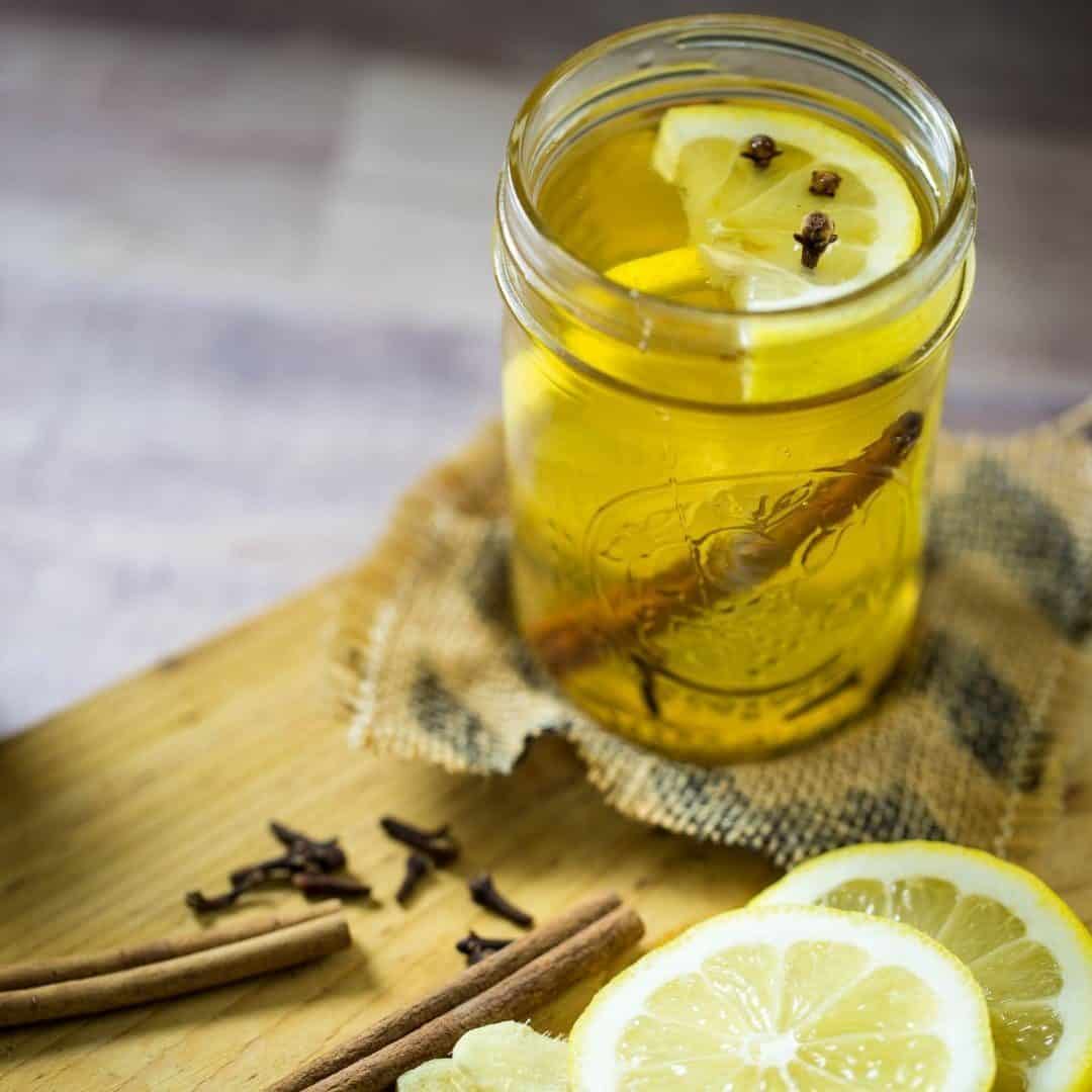 A mason jar filled with a homemade hot toddy garnished with lemon wedge whole cloves and a cinnamon stick.