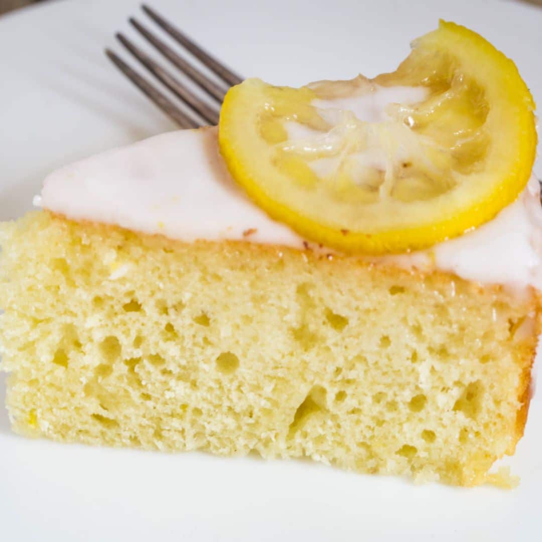 A slice of lemon yogurt cake on a white plate with a lemon wedge on top of the piece of cake.