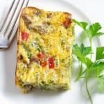 A delicious egg bake made with eggs, sausage, broccoli, peppers, and cheese on a white plate with a fork and a sprig of parsley on the plate.