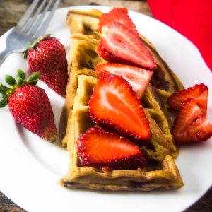 Homemade strawberry waffles stacked on a white plate with fresh strawberries on top of the waffles.