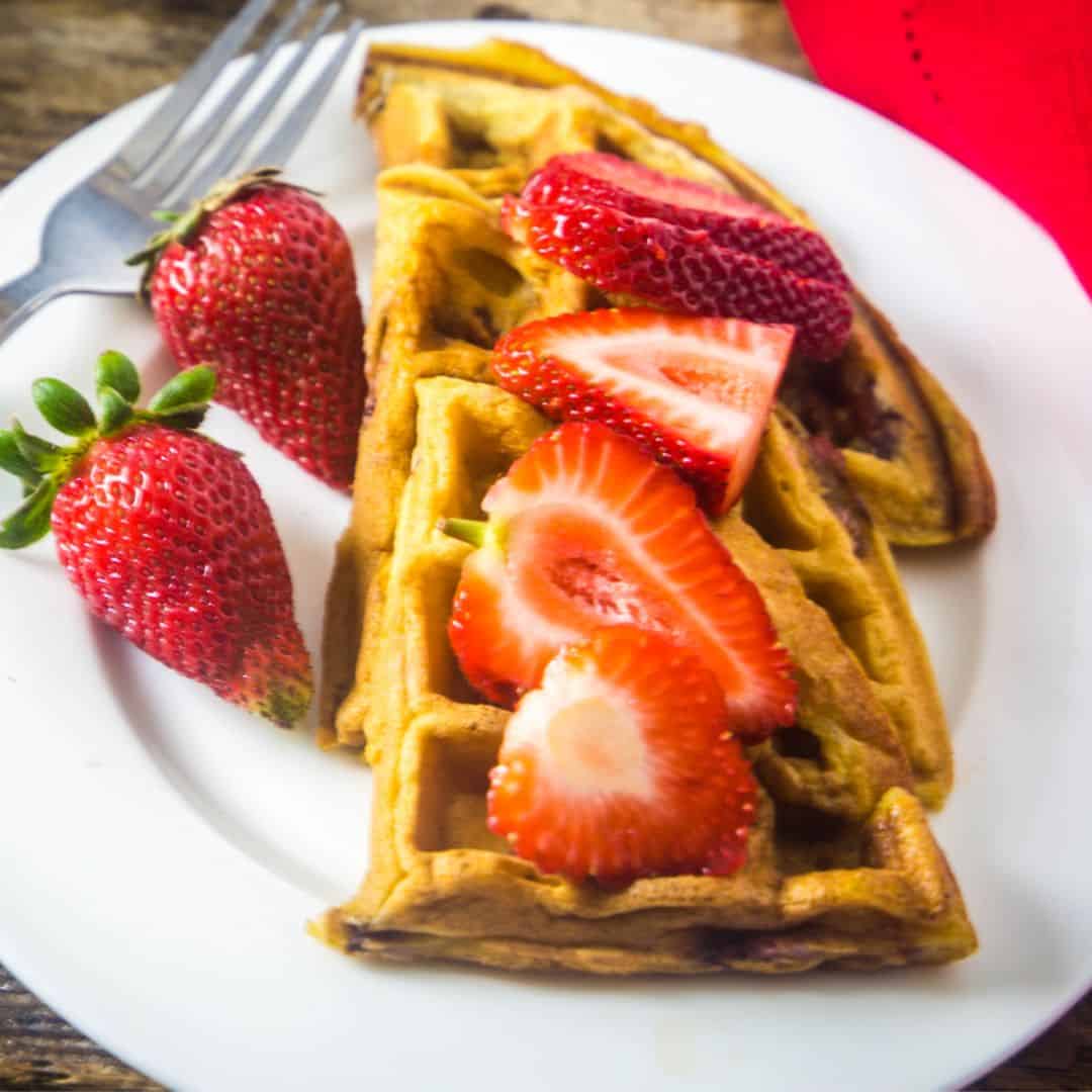 Strawberry waffles on a white plate with fresh strawberries drizzled over the waffles.