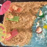 Festive beach cake decorated with blue frosting and graham cracker sand with teddy graham bears on the beach under beach umbrellas