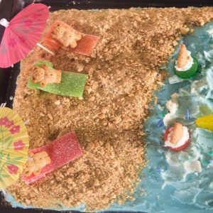 Festive beach cake decorated with blue frosting and graham cracker sand with teddy graham bears on the beach under beach umbrellas