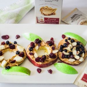 Apple slices topped with peanut butter, almonds, dried cranberries on a white platter