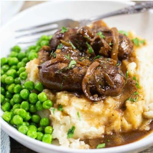 A mound of mashed potatoes topped with sausages and caramelized onions with green peas on the side