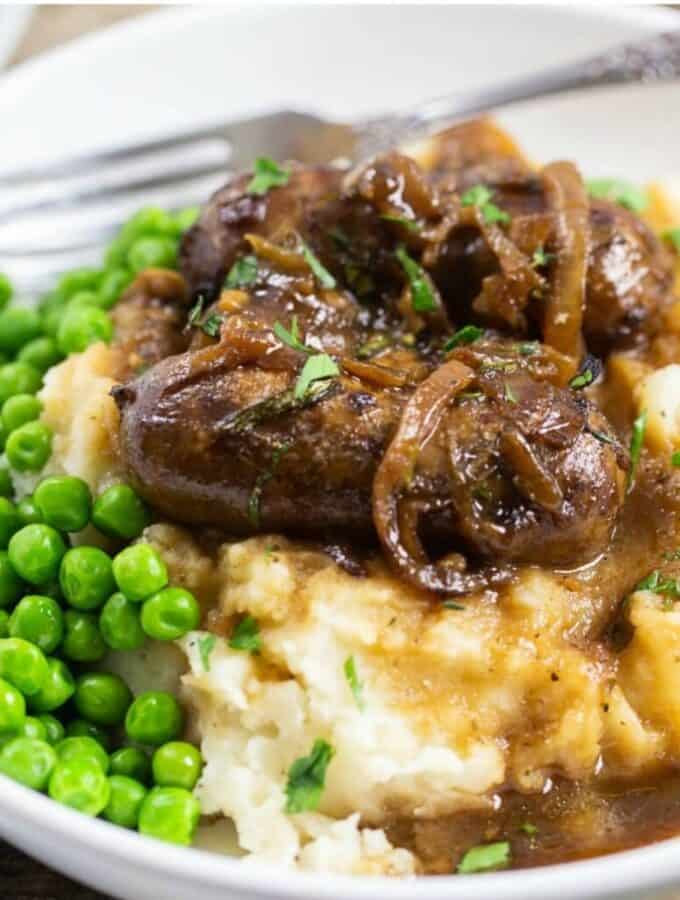 A mound of mashed potatoes topped with sausages and caramelized onions with green peas on the side