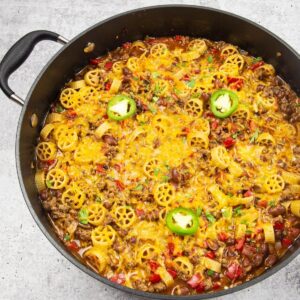 A large pan filled with spicy ground beef and wagon wheel pasta topped with melted cheese and sliced jalapeno