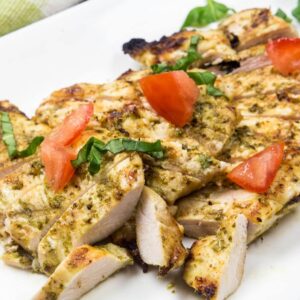 Sliced boneless chicken breasts coated with basil pesto on a white plate, topped with chopped fresh basil and tomato.