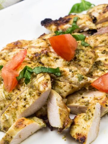 Sliced boneless chicken breasts coated with basil pesto on a white plate, topped with chopped fresh basil and tomato.