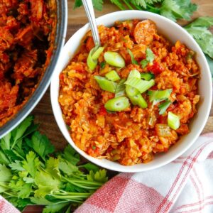 A white bowl filled with jambalaya and cauliflower rice topped with chopped green onions and a bunch of fresh parsley on the side.