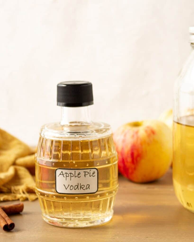 A homemade bottle of apple pie vodka with fresh apples and cinnamon on the side.
