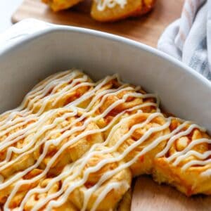 A white casserole dish with baked pumpkin flavored cinnamon rolls topped with cream cheese frosting