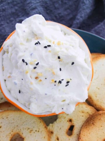 A creamy dip in a bowl topped with everything but the bagel seasoning and surrounded with bagel chips