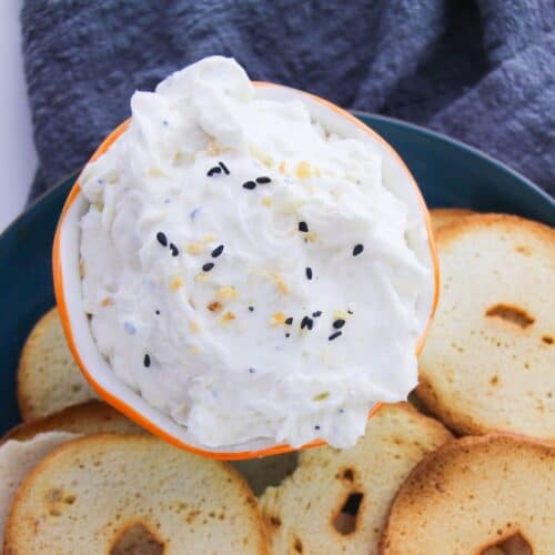 A creamy dip in a bowl topped with everything but the bagel seasoning and surrounded with bagel chips